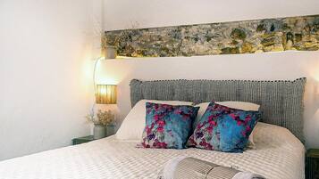 Una cama con edredón blanco texturizado, dos almohadas decorativas con motivos florales, un cabecero con diseño texturizado y una lámpara de pared sobre una mesita de noche.