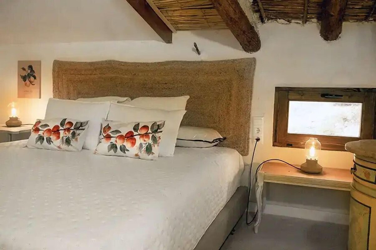 Una cama con almohadas estampadas, una mesita de noche con lámpara y una ventana con vistas.