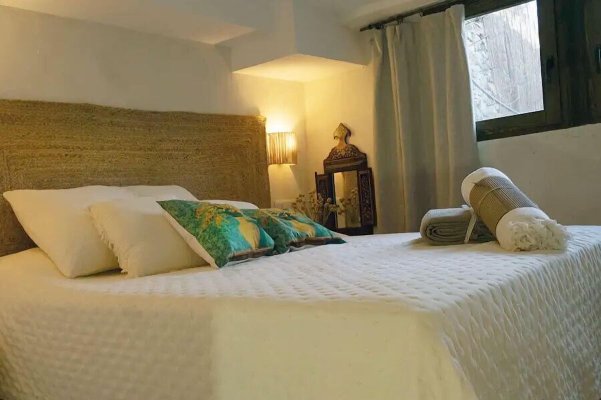 Una cama con ropa blanca y almohadas decorativas, un cabecero, una lámpara de noche y una ventana con cortinas.