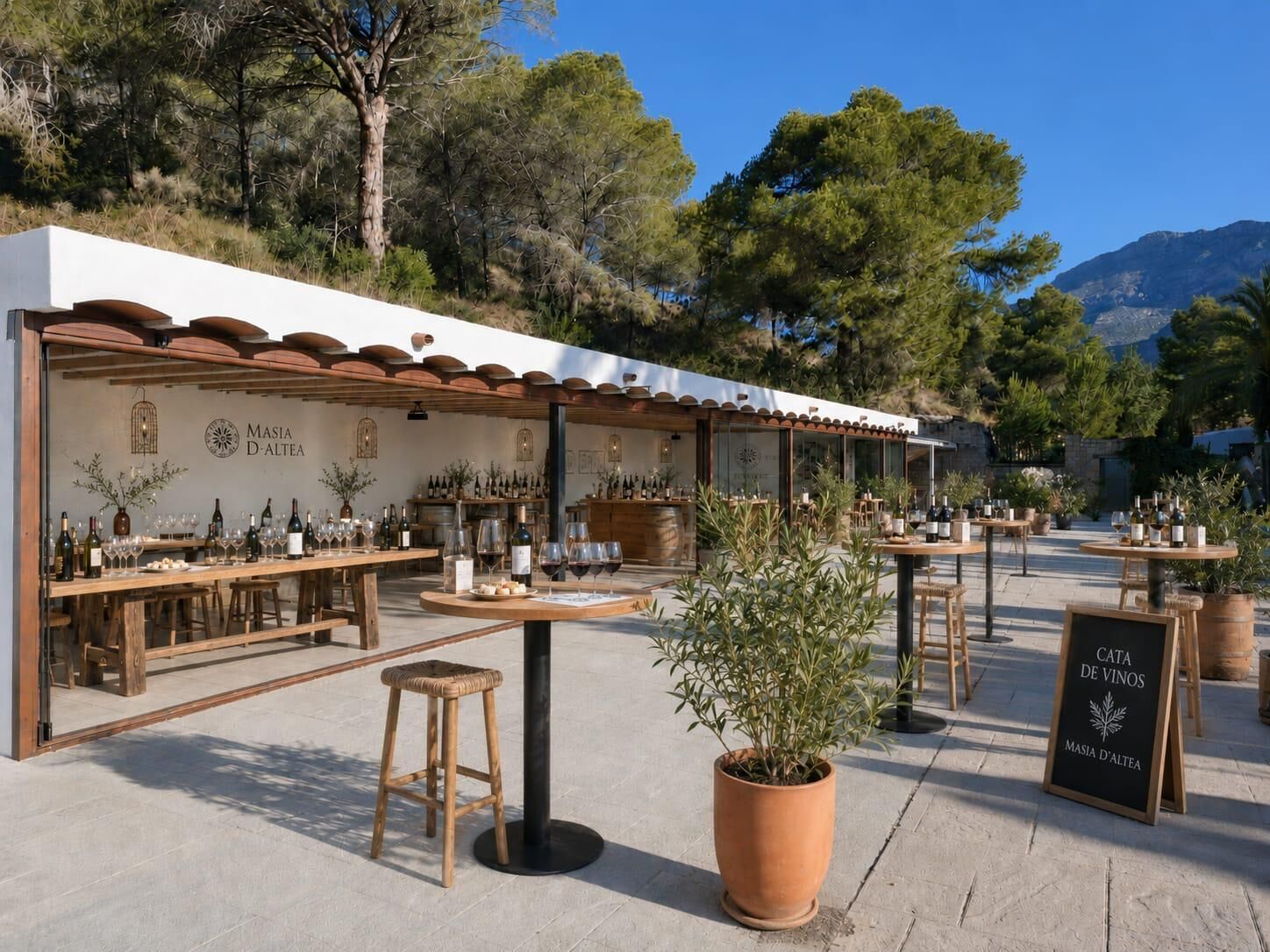 An outdoor seating area with tables and chairs, surrounded by potted plants and a sign for 'CATA DE VINOS' at Masia D'Altea.