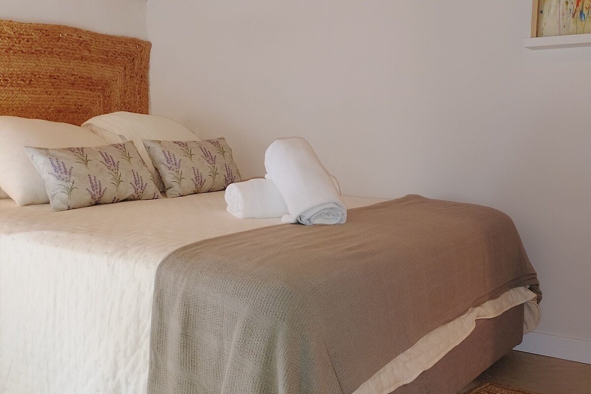Una cama bien hecha con una colcha beige y blanca, una toalla blanca enrollada y dos almohadas con estampado floral.