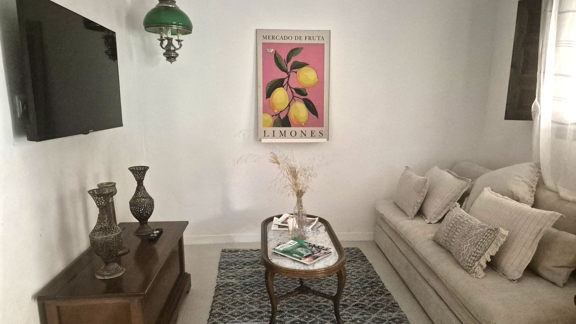 Una sala de estar con sofá, una mesa de centro con revistas, un televisor de pared, un cartel decorativo y una lámpara de pared verde.