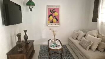 Una sala de estar con sofá, una mesa de centro con revistas, un televisor de pared, un cartel decorativo y una lámpara de pared verde.
