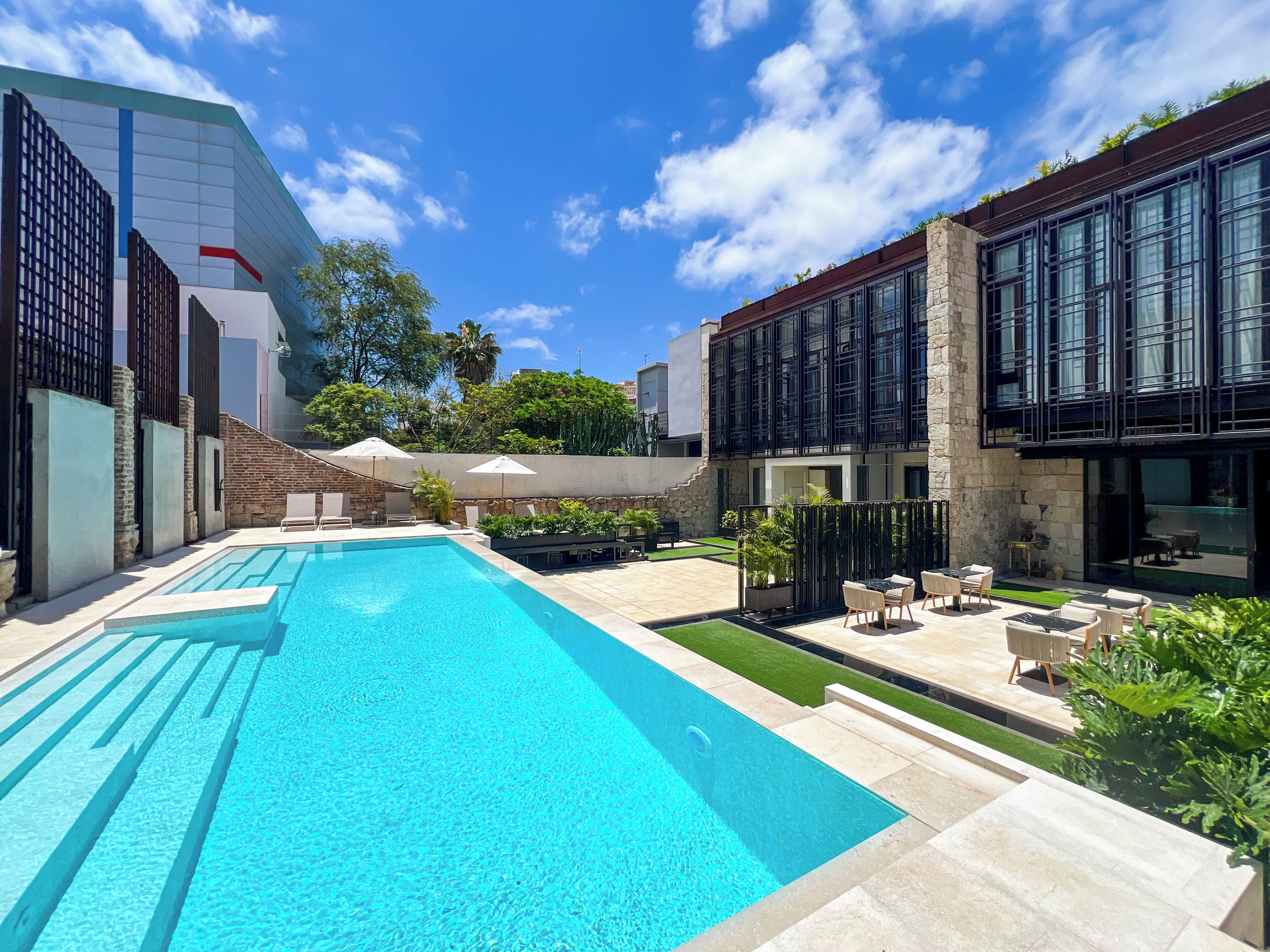 A swimming pool with lounge chairs and umbrellas, surrounded by modern buildings and greenery.