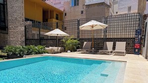 A swimming pool with lounge chairs and umbrellas, surrounded by a stone wall and a metal fence.