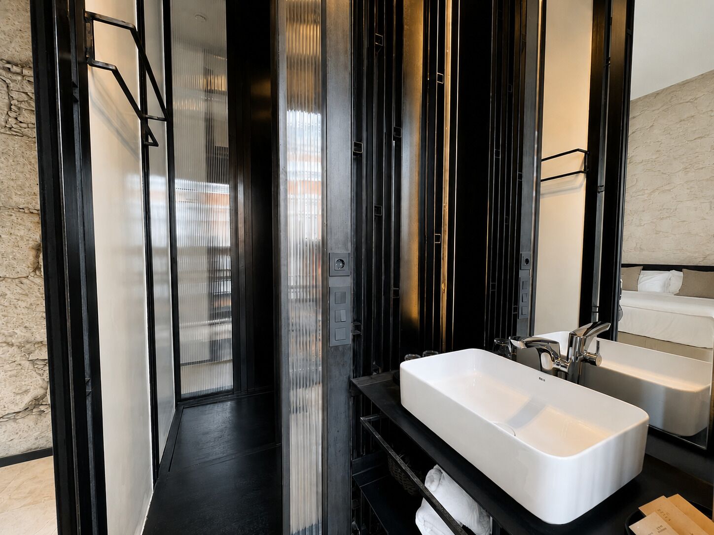 A modern bathroom with a large white sink, black shelves, and a glass door leading to a shower area.