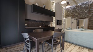 Una cocina moderna con armarios oscuros, una mesa de comedor y sillas. Se observa una pared de piedra y una cama con colchón blanco y ropa de cama azul.