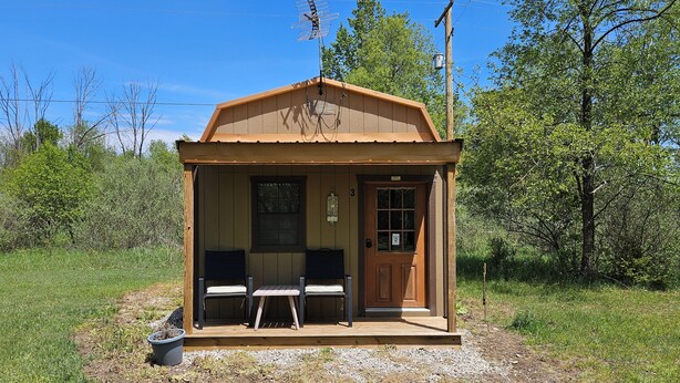 Een klein houten vakantiehuisje met een veranda, twee stoelen, een tafel en een deur.