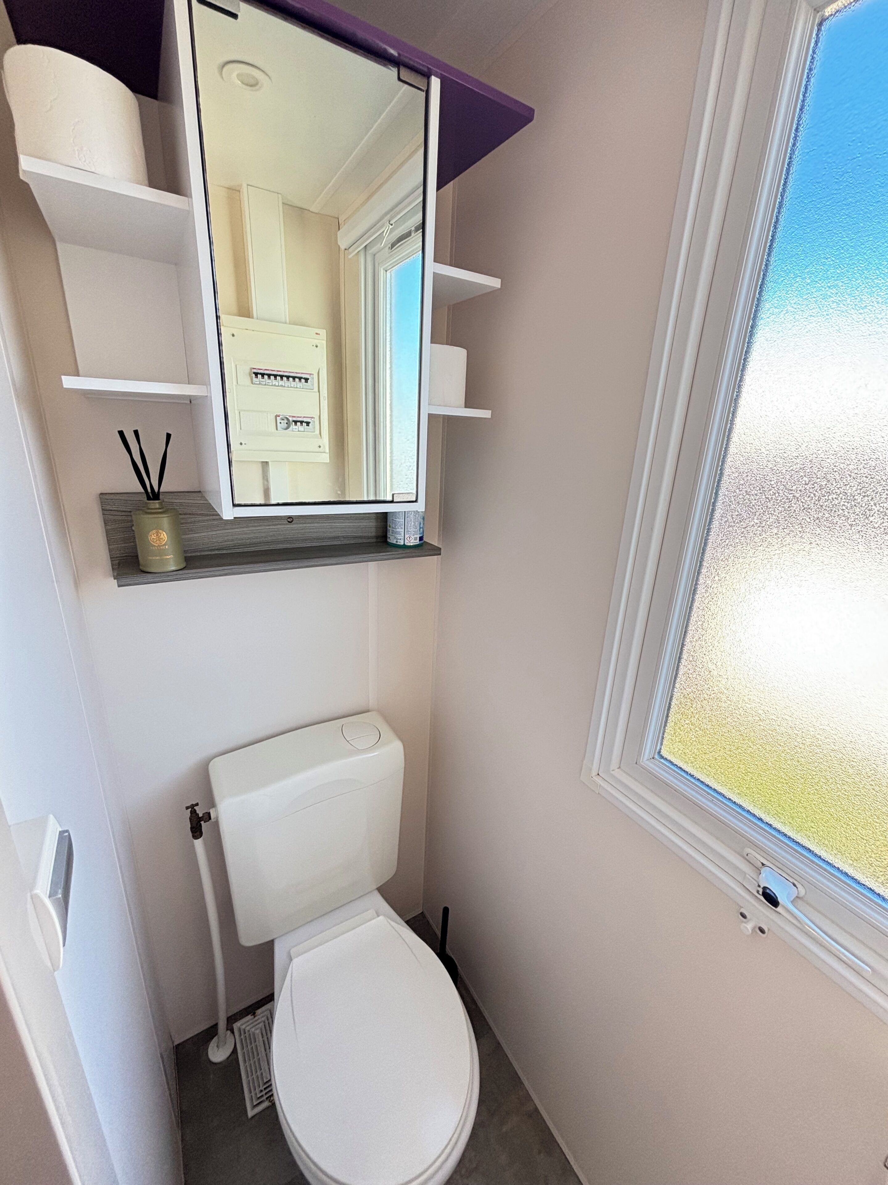 Een kleine badkamer met toilet, spiegel, raam en een plank met toiletartikelen.