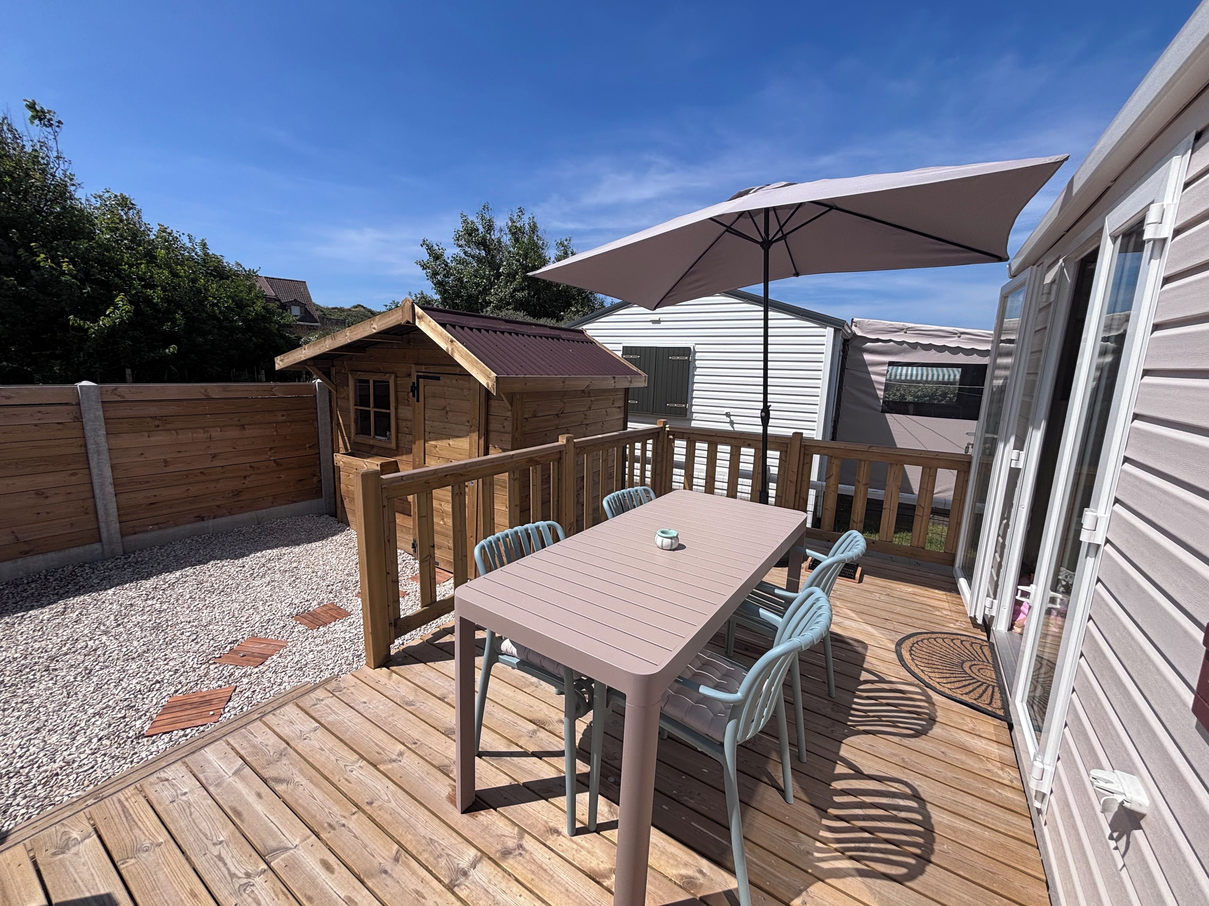 Een houten terras met een tafel, stoelen en een grote parasol, plus een klein houten bouwwerk.