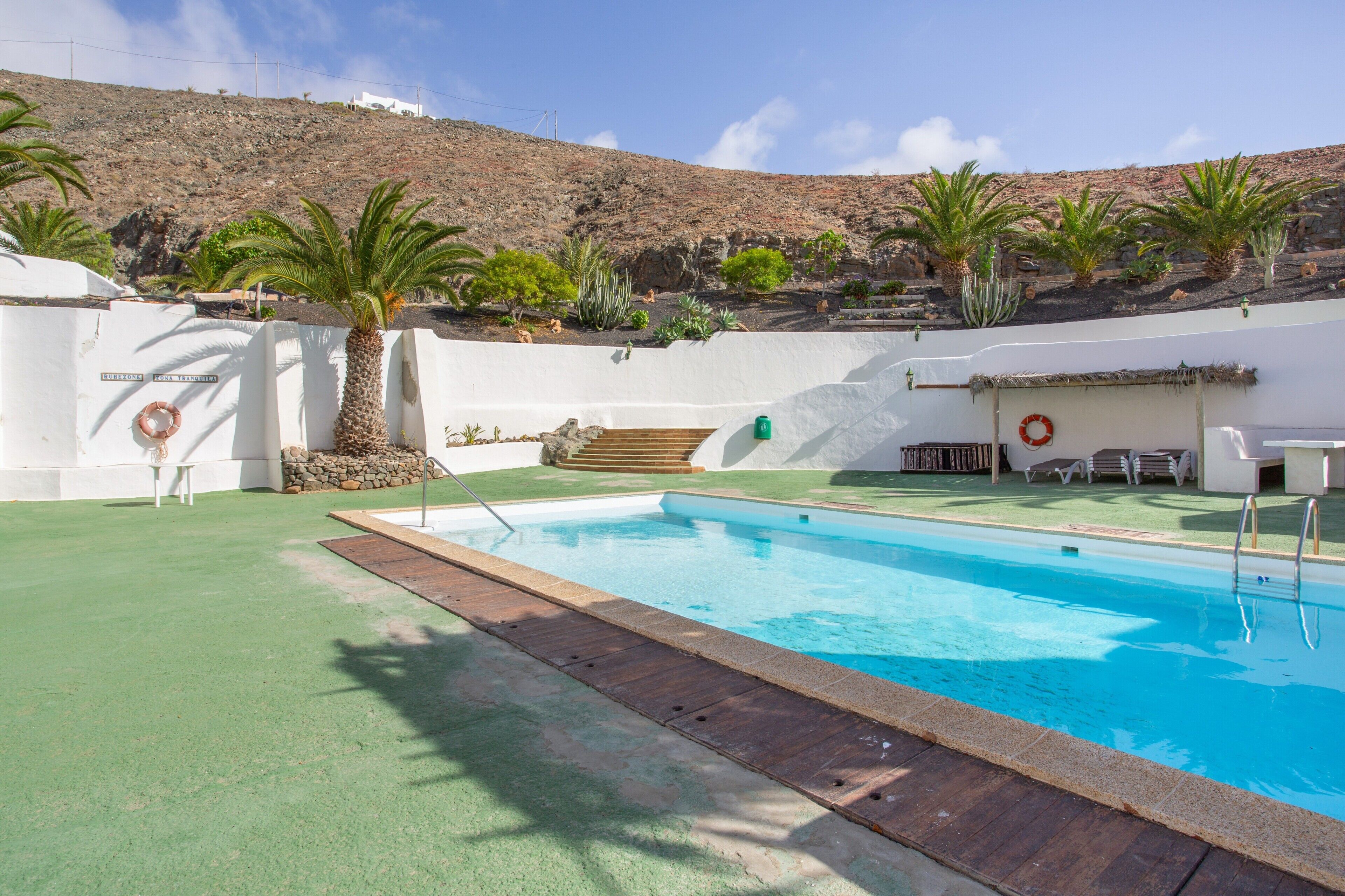 Une piscine avec une terrasse en bois, des palmiers et un mur blanc orné d’une bouée de sauvetage.