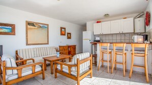 A living room with a sofa, wooden chairs, a coffee table, and a kitchen area with a refrigerator, stove, and bar stools. - Apartment in a Natural Park on Fuerteventura’s West Coast (Valle de Santa Inés)