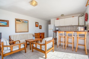 A living room with a sofa, wooden chairs, a coffee table, and a kitchen area with a refrigerator, stove, and bar stools. - Apartment in a Natural Park on Fuerteventura’s West Coast (Valle de Santa Inés)