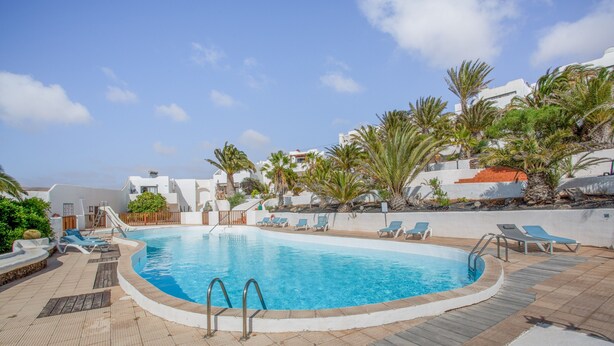 A swimming pool with lounge chairs, palm trees, and a white building in the background. - Apartment in a Natural Park on Fuerteventura’s West Coast (Valle de Santa Inés)