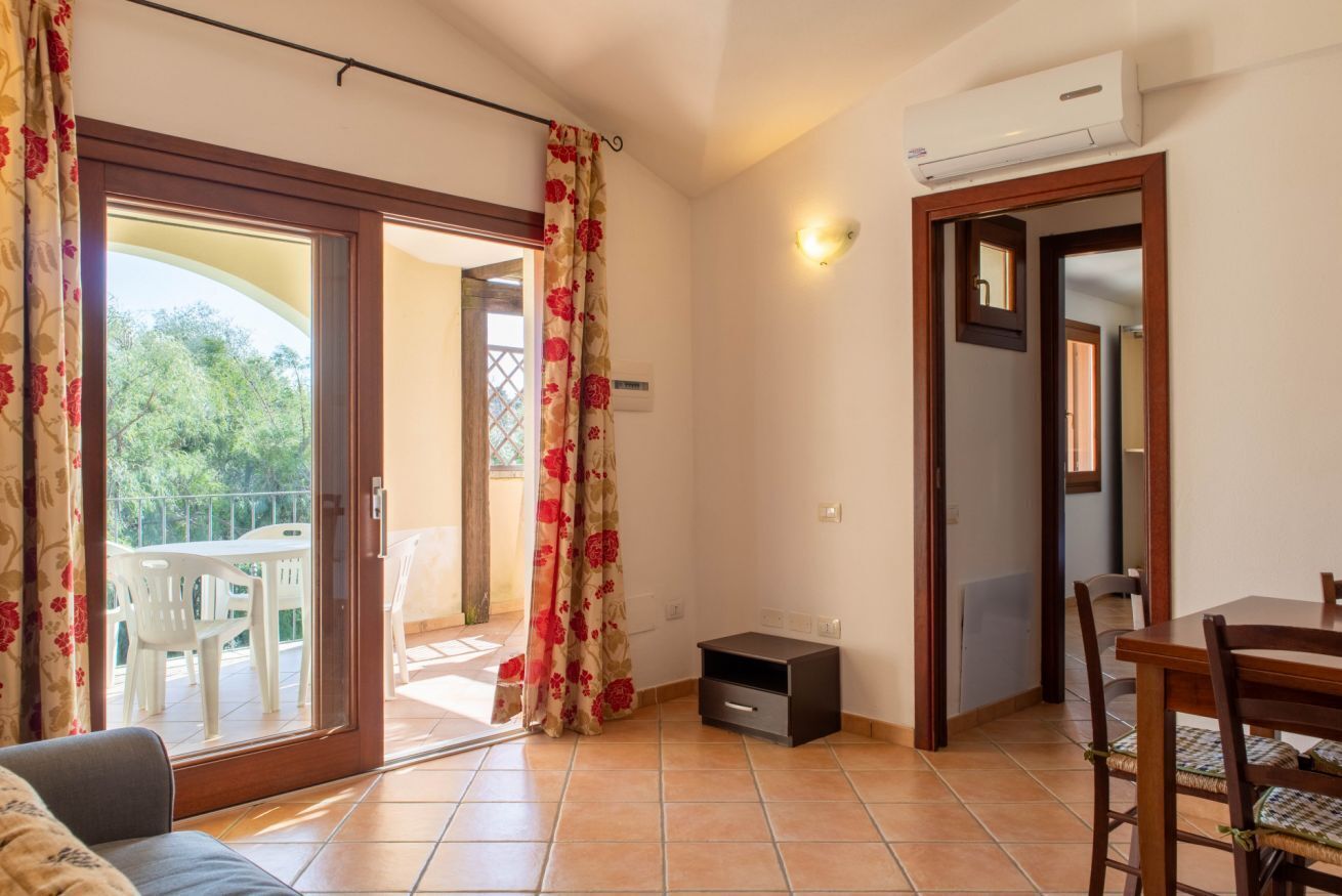 A room with a sliding glass door leading to a balcony, a small table with chairs, a sofa, and an air conditioning unit mounted on the wall.