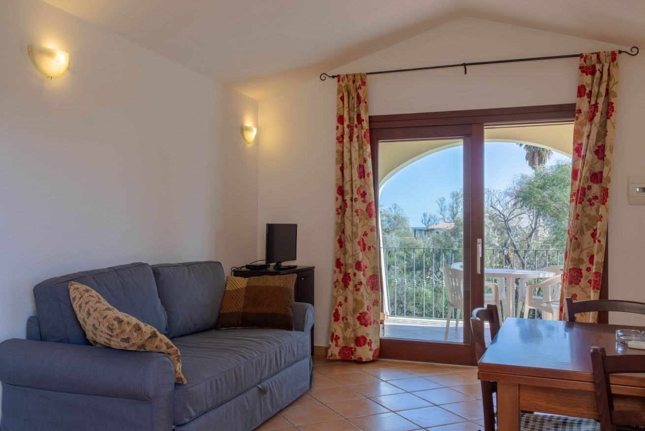 Ein Wohnzimmer mit Sofa, Fernseher auf einem Schrank, einem Esstisch mit Stühlen und einem Balkon mit Aussicht.