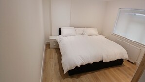Un dormitorio con una cama, una mesita de noche, un ventanal con persianas y suelo de madera.