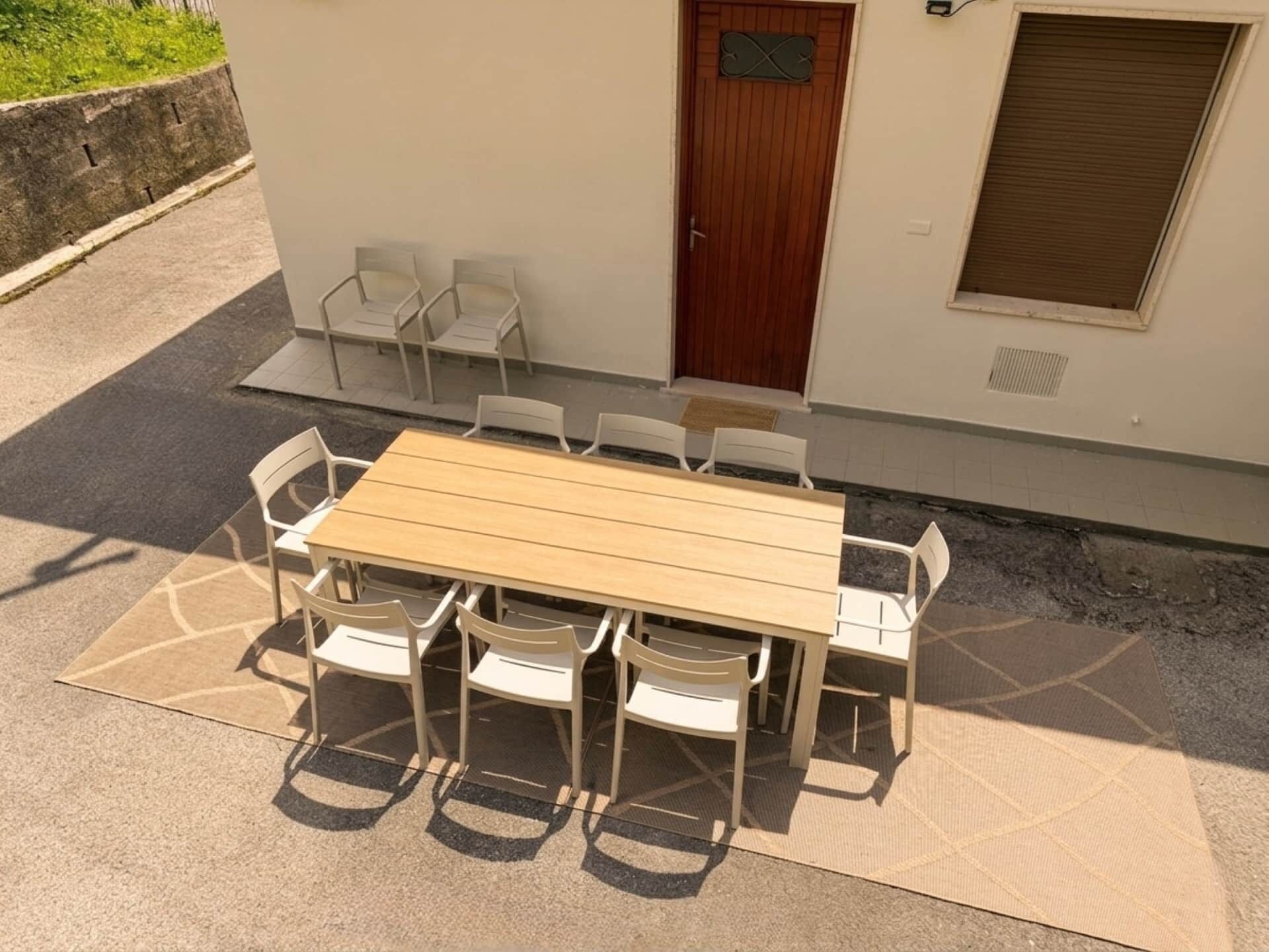 Een buitenterras met een houten tafel en witte stoelen, een deur en een raam met een luik.