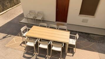 Een buitenterras met een houten tafel en witte stoelen, een deur en een raam met een luik.