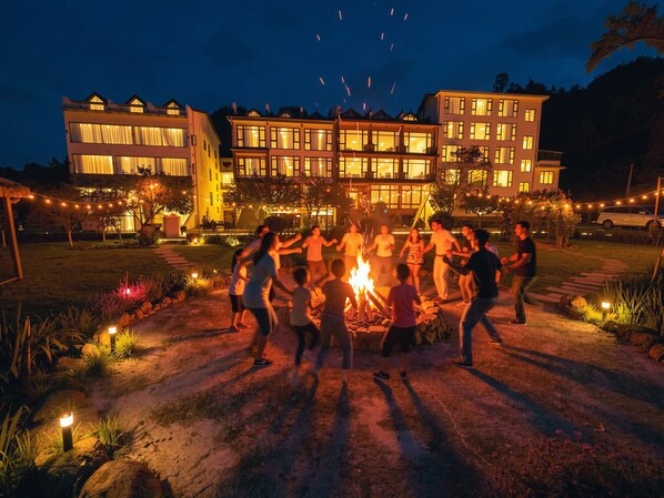 Eine Gruppe von Menschen tanzt um ein Lagerfeuer in einer Freifläche, im Hintergrund ist ein Gebäude zu sehen.