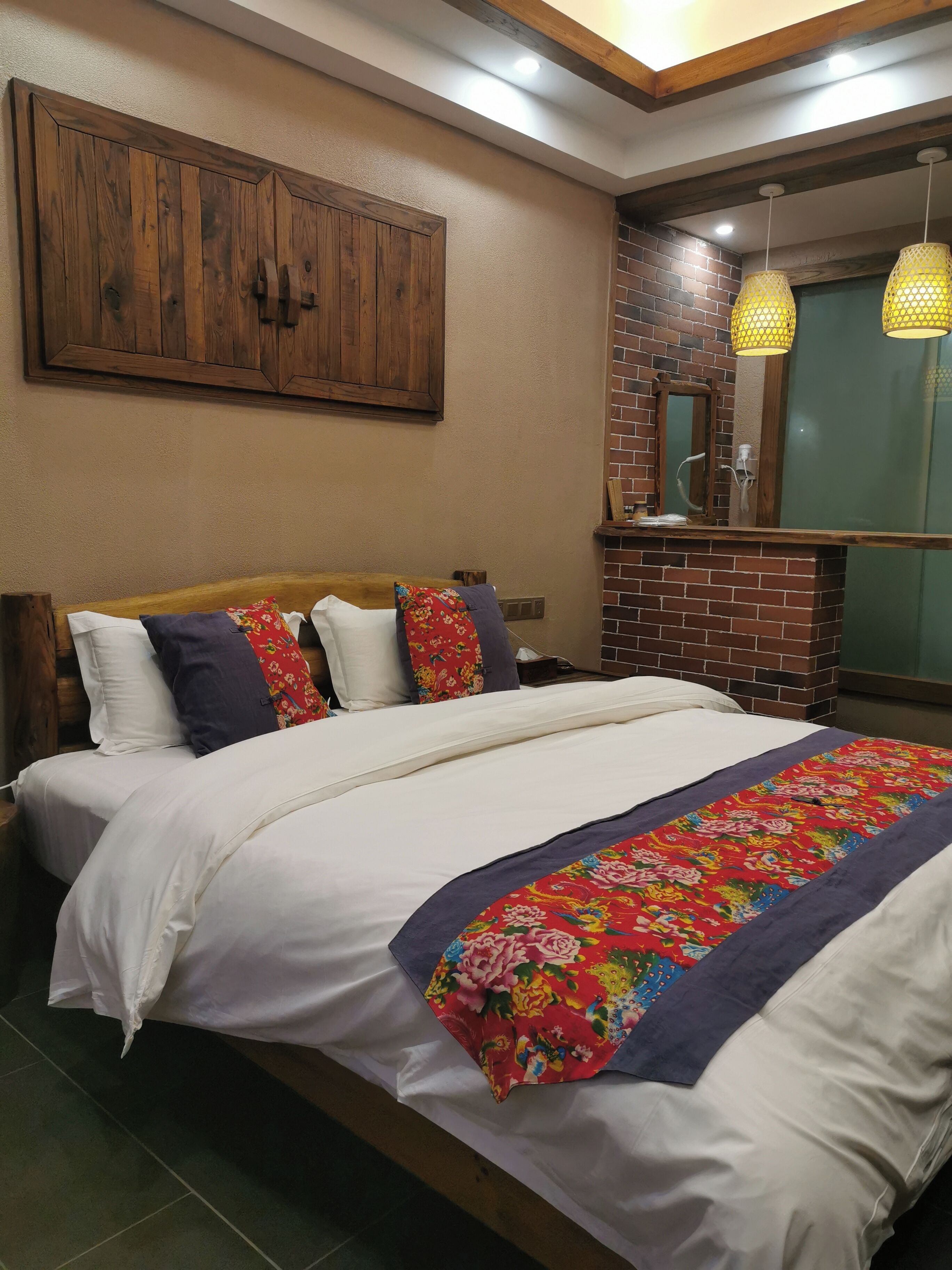 A bedroom with a wooden headboard, a bed with white bedding and a floral throw, a brick accent wall, and a hanging light fixture.