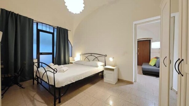 A bedroom with a bed, bedside table, and a view of a living area through an open door. - Hostdomus - Oscar's House (Albenga)