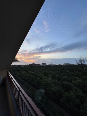 Ein Balkon mit Blick auf einen Sonnenuntergang über einer Landschaft mit spärlicher Vegetation und weit entfernten Gebäuden.