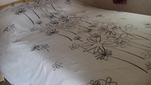 En seng med et blomstret sengetæppe, en træhylde over og et vindue med gardin.