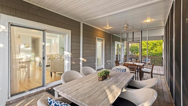 Eine überdachte Terrasse mit Holztisch, Korbstühlen und einer Glasschiebetür zu einem Innenbereich mit Sofa und Sesseln.