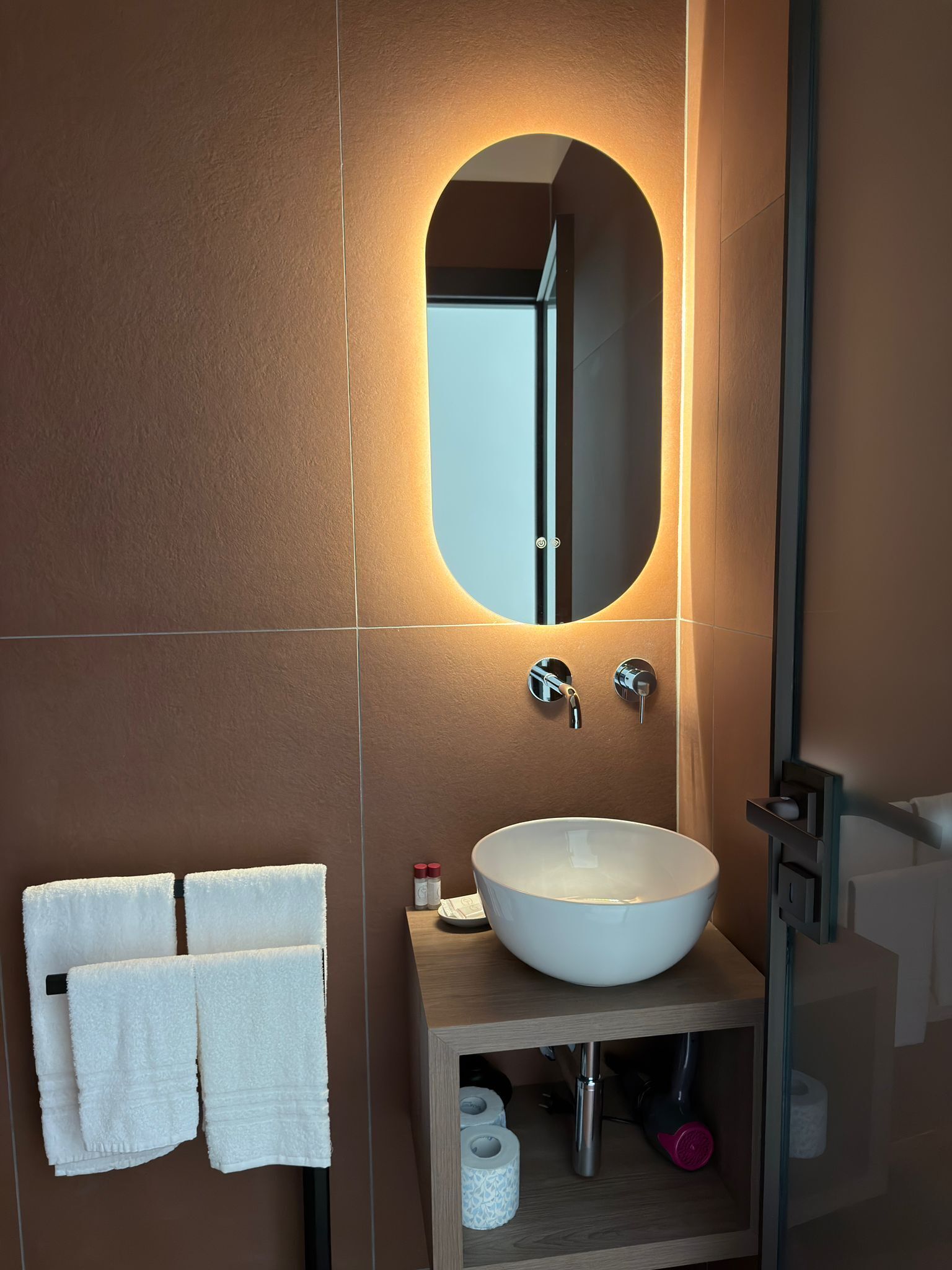 A bathroom with a large oval mirror, a white sink, two faucets, a towel rack with two towels, and a small shelf with toiletries.