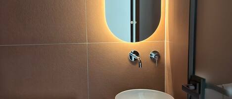 A bathroom with a large oval mirror, a white sink, two faucets, a towel rack with two towels, and a small shelf with toiletries.
