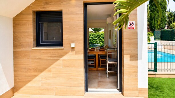 Een modern gebouw met een houten gevel, een zichtbare eetruimte door een geopende deur en een zwembad op de achtergrond.