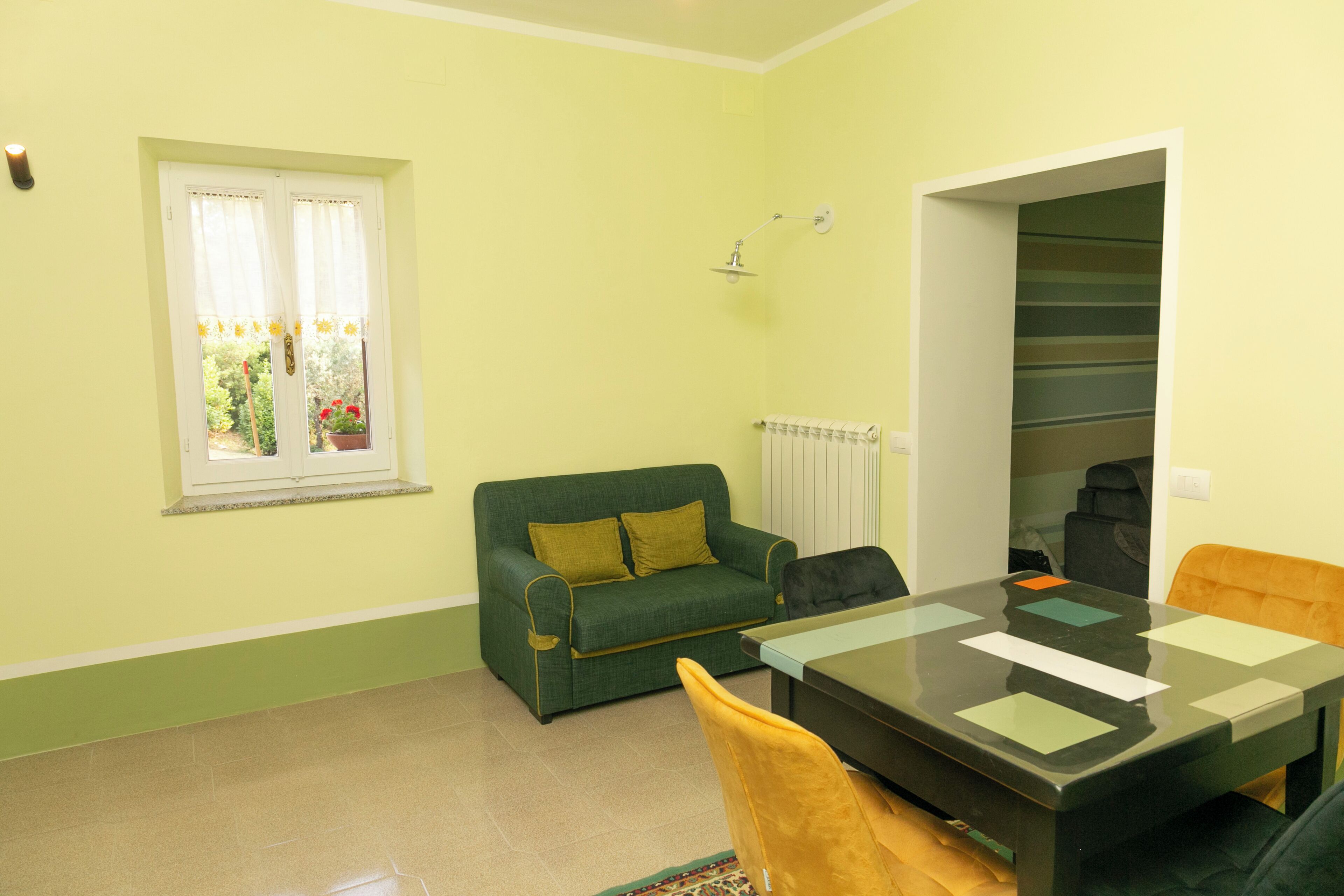 Een kamer met een groene bank, een eettafel, een stoel, een raam met gordijnen en een radiator.