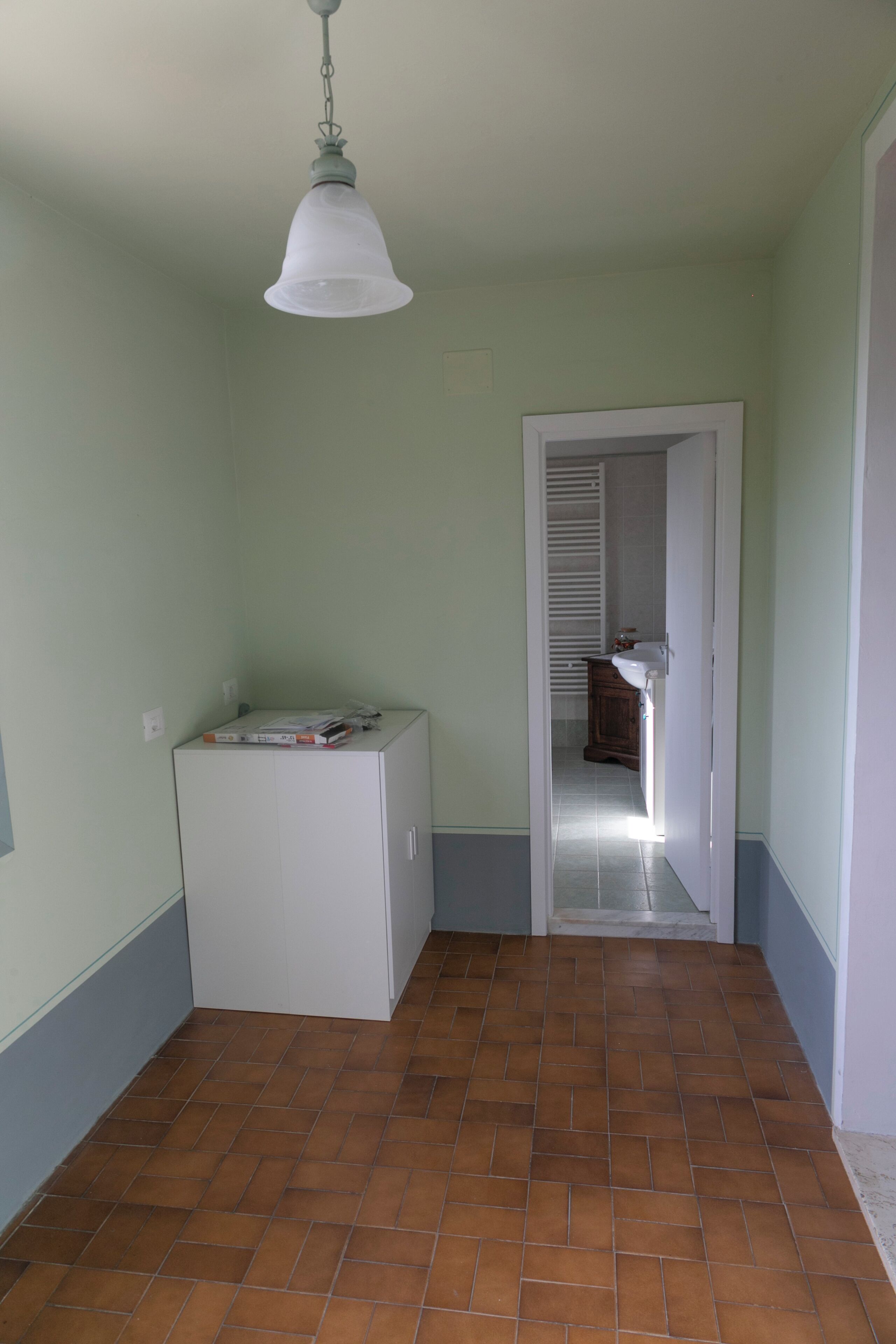 A hallway with a white cabinet, a hanging light fixture, and a door leading to a bathroom.
