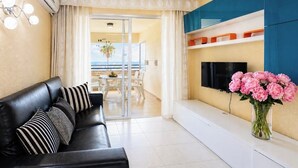 Ein Wohnzimmer mit einer schwarzen Ledersofa, einem Flachbildfernseher, einer Blumenvase und Blick durch eine Glastür auf einen Balkon.