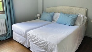 Cama de casal com roupa de cama branca e duas almofadas azuis, mesa de cabeceira com luminária e janela com cortinas azul-claras.