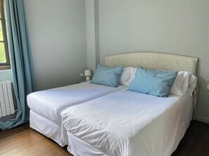 A double bed with white bedding and two blue pillows, a nightstand with a lamp, and a window with light blue curtains. - Charming Rural Retreat with Garden — Countryside & near the Coast for up to 6 (Reocín)