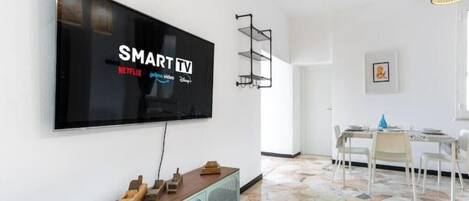 A modern living room with a large flat-screen TV mounted on the wall, a dining area with a table and chairs, and a patterned floor.