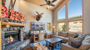 Un salon chaleureux avec une cheminée en pierre, une tête de cerf accrochée au mur et une grande fenêtre donnant sur le paysage extérieur.