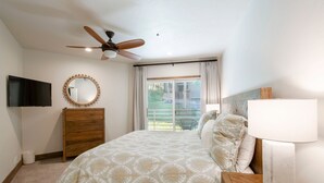 Een slaapkamer met een bed, een plafondventilator, een televisie, een dressoir, een lamp en een raam met gordijnen.