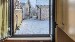 Ein Zimmer mit großem Fenster, Holzwänden, integrierter Heizung und Blick auf einen verschneiten Innenhof mit Häusern.