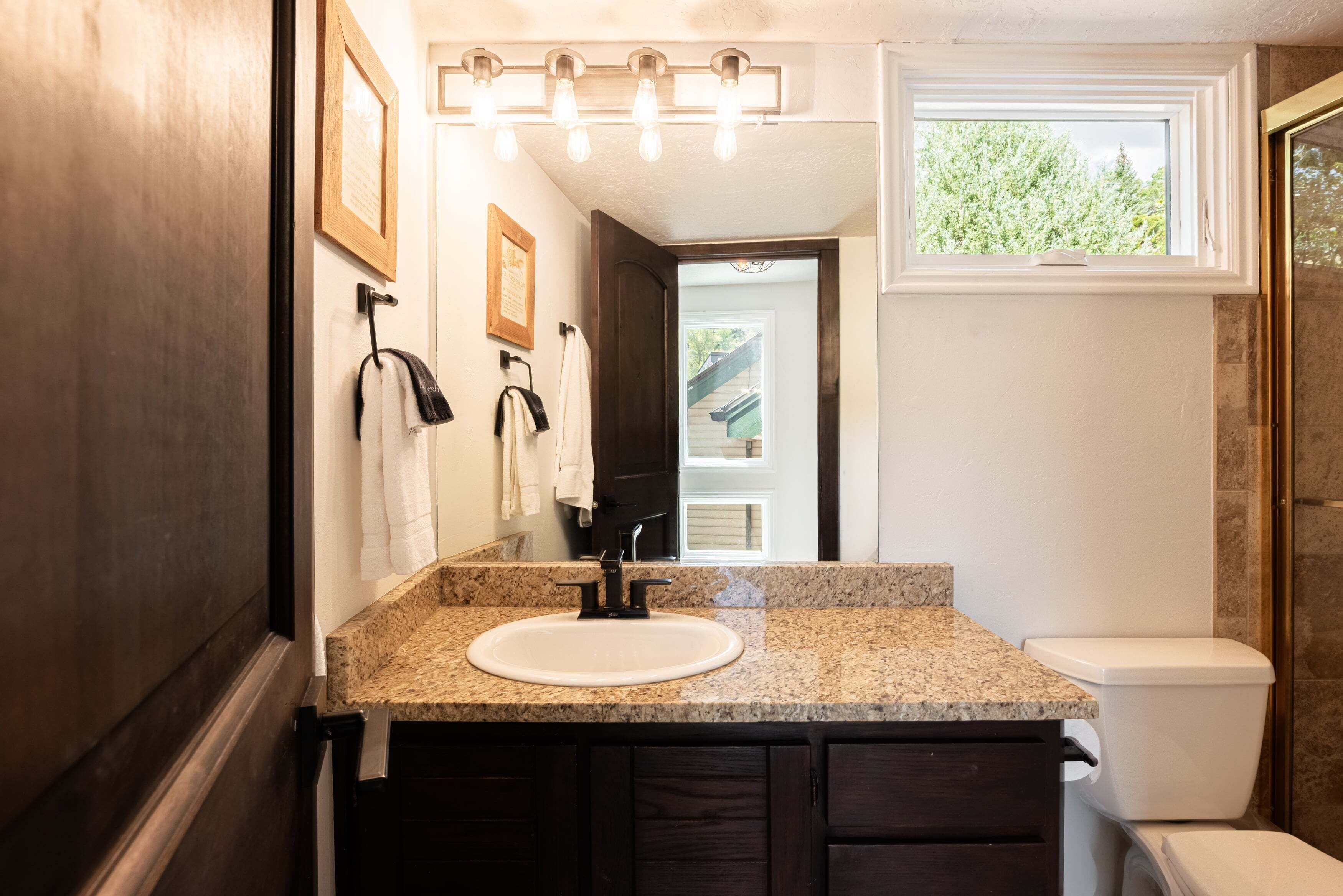 Une salle de bain dotée d’un comptoir en granit, d’un lavabo blanc, d’un robinet noir, d’armoires en bois foncé, de toilettes blanches et d’une fenêtre donnant sur un paysage verdoyant.