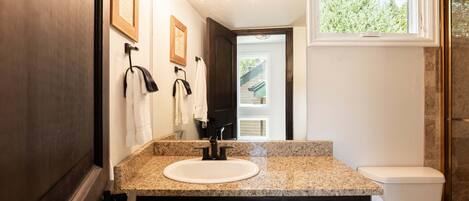 Une salle de bain dotée d’un comptoir en granit, d’un lavabo blanc, d’un robinet noir, d’armoires en bois foncé, de toilettes blanches et d’une fenêtre donnant sur un paysage verdoyant.