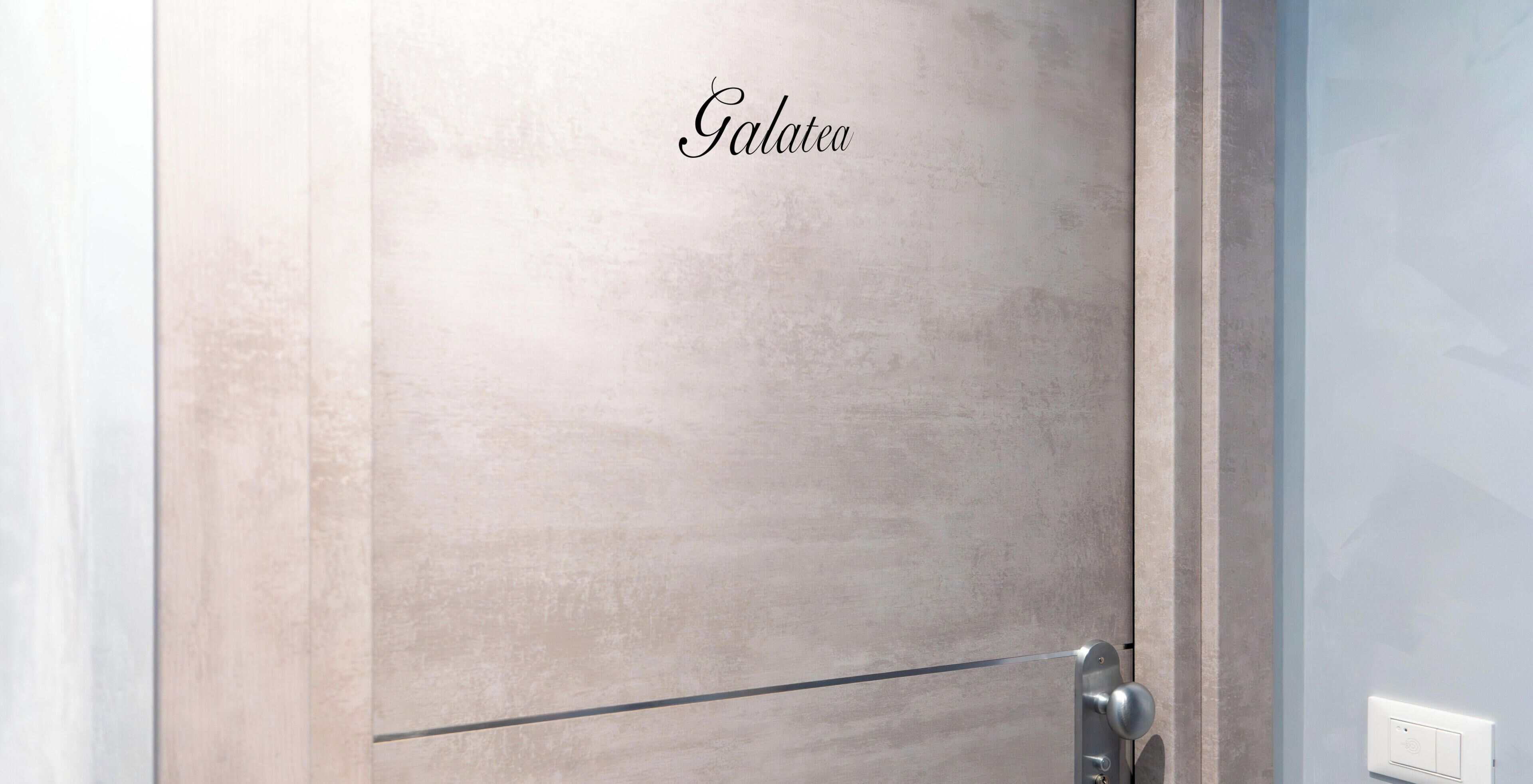 A door with the word 'Galatea' on it, a metallic handle, and a light switch on the wall.