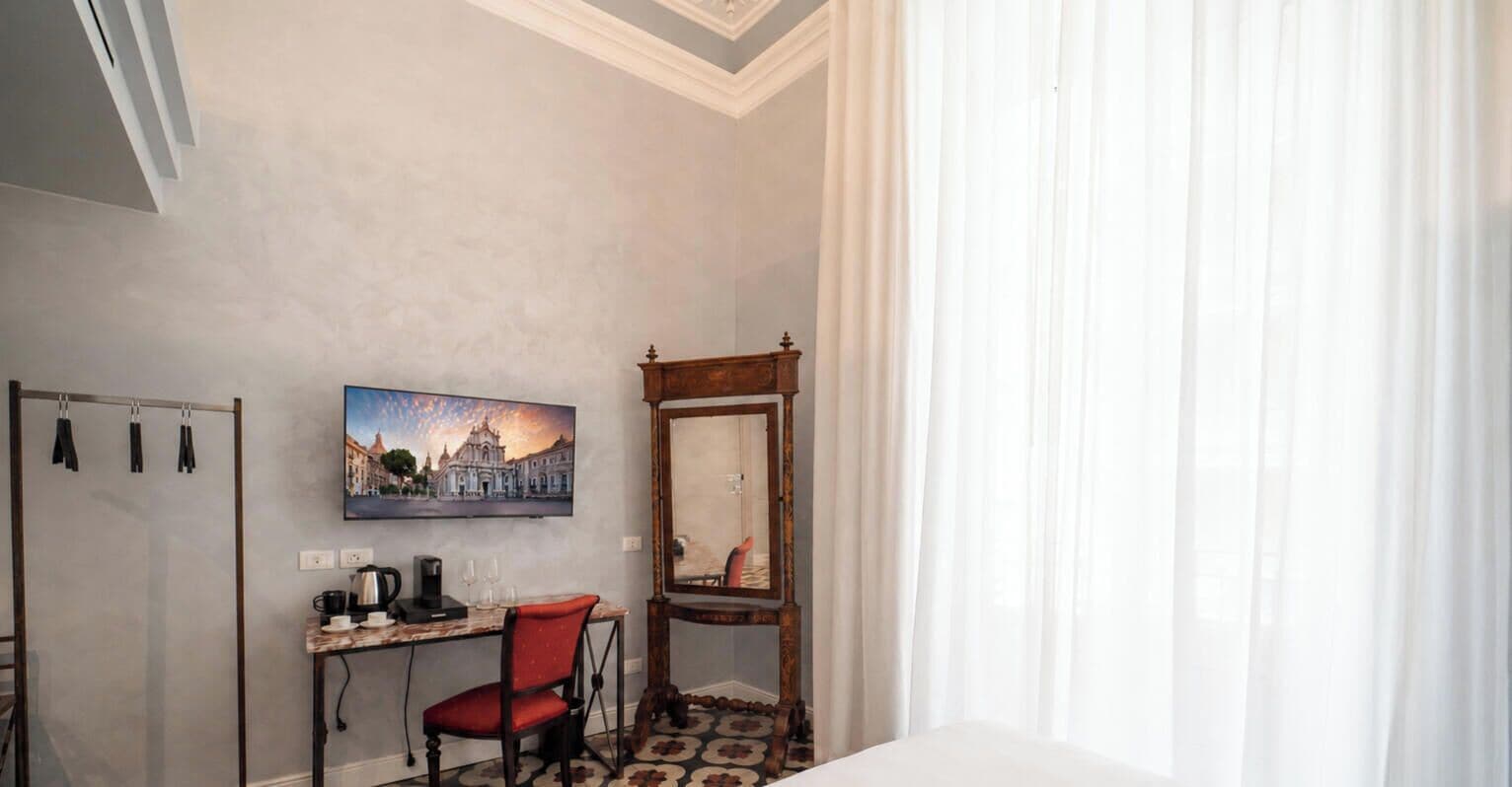 A hotel room with a bed, a desk with a chair, a painting, a mirror, and a window with curtains.