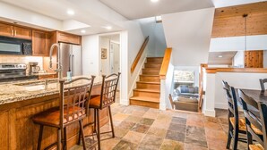 Eine Küche mit einer zentralen Kochinsel, Holzschränken, einem Edelstahlkühlschrank und einer Treppe, die zu einem Erkerraum führt.