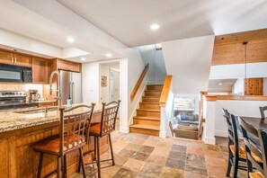 Eine Küche mit einer zentralen Kochinsel, Holzschränken, einem Edelstahlkühlschrank und einer Treppe, die zu einem Erkerraum führt.