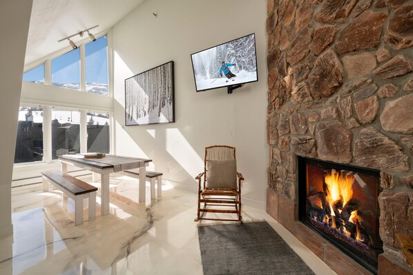 A modern living room with a stone fireplace, a mounted TV, and a large window overlooking a snowy landscape. - Ski-In/Out Base Village | 3BR w/ Pool, Gym & Hot Tub (Park City)