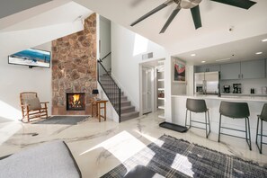 A modern living room with a stone fireplace, a TV mounted on the wall, a wooden rocking chair, and a staircase leading to another level. - Ski-In/Out Base Village | 3BR w/ Pool, Gym & Hot Tub (Park City)