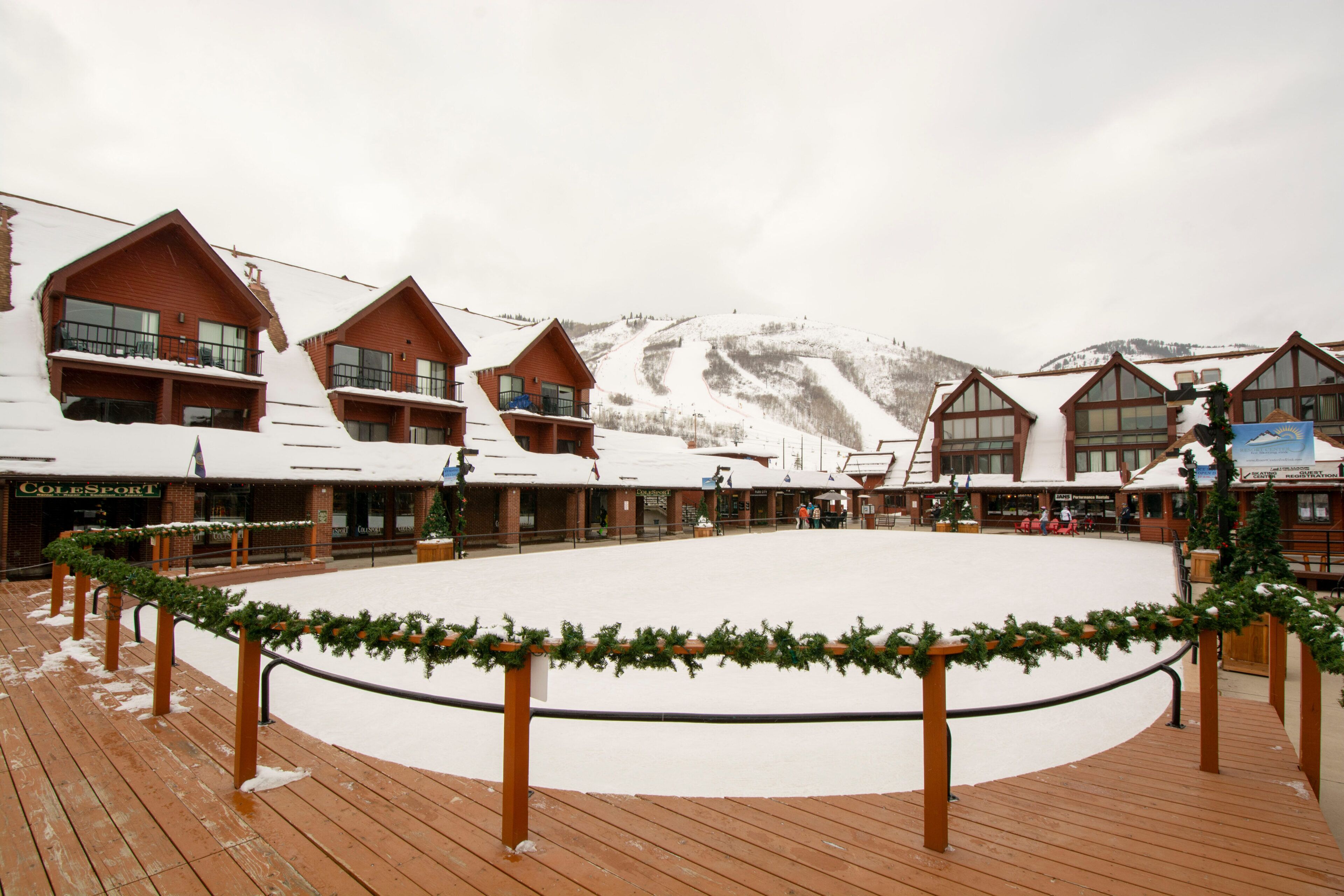 Una terraza de madera cubierta de nieve con un área de estar circular, rodeada de edificios con grandes ventanales y un paisaje montañoso de fondo.